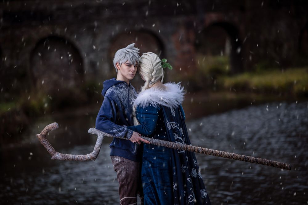 Zwei Personen in winterlichen Fantasy-Kostümen stehen sich nahe an einem nebligen Flussufer gegenüber. Eine Figur hat silbernes, verwuscheltes Haar und trägt einen dunkelblauen Hoodie mit frostigem Muster sowie eine braune Hose. In der Hand hält sie einen langen, geschwungenen Stab, der mit Stoff umwickelt ist. Die andere Person hat langes, blondes Haar, kunstvoll zu einem Zopf geflochten, und trägt einen eleganten, dunkelblauen Mantel mit weißen Schneeflockenmustern und einem flauschigen Kragen. Die Szene ist umgeben von sanft fallenden Schneeflocken, während im Hintergrund eine alte Steinbrücke zu sehen ist. Die Stimmung ist magisch und geheimnisvoll.