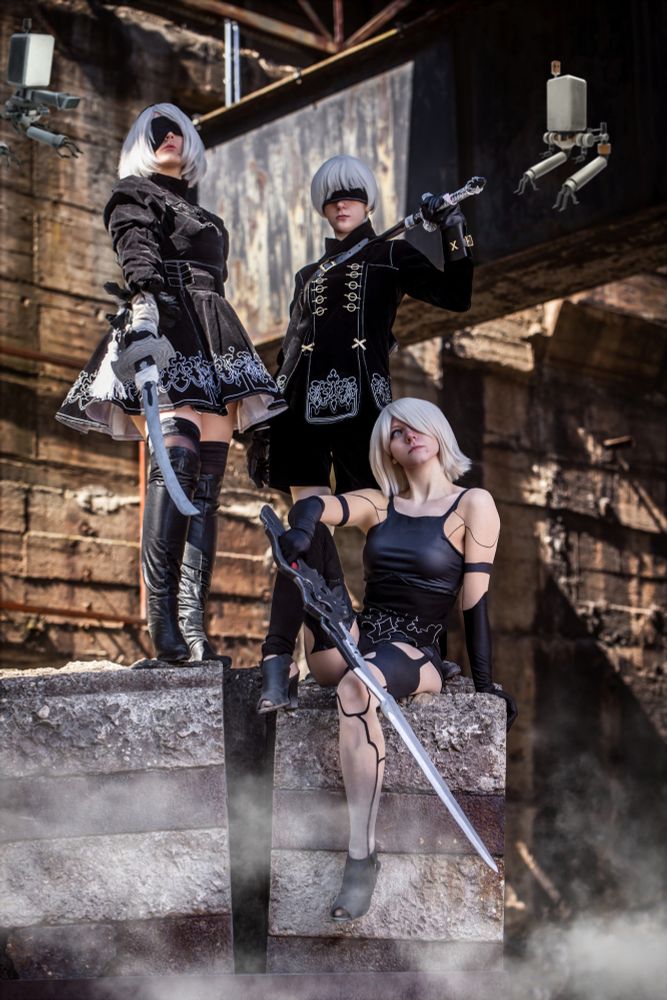 Drei Cosplayerinnen stellen Charaktere aus dem Spiel „NieR:Automata“ dar. Sie posieren in einem verlassenen Industriekomplex auf massiven Betonblöcken. Links steht eine Person als 2B in einem schwarzen Kleid mit weißen Stickereien und kniehohen Stiefeln, das Gesicht teilweise von einer weißen Perücke und einer schwarzen Augenbinde verdeckt. Rechts daneben steht eine zweite Person als 9S, ebenfalls mit Augenbinde und schwarzem, uniformähnlichem Outfit. In der Mitte sitzt eine dritte Person als A2 auf einem Betonblock, mit kurzem Kleid, Armstulpen und auffälligem Schwert. Über ihnen schweben zwei kleine Roboter-Drohnen, sogenannte Pods. Dampf steigt vom Boden auf und verstärkt die dystopische Stimmung.