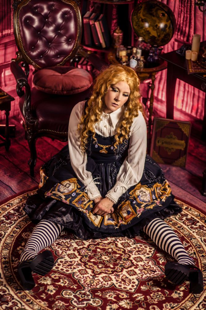 Eine Person mit langen, goldblonden Locken und nostalgisch wirkendem Make-up sitzt wie eine leblose Porzellanpuppe auf einem orientalischen Teppich in einem viktorianisch eingerichteten Raum. Sie trägt ein Lolita-Kleid mit barocken Porträtmotiven, eine weiße Bluse mit Rüschen und schwarz-weiß gestreifte Strumpfhosen. Die Umgebung ist reich verziert mit antiken Möbeln, Lederstuhl, Globus, Kerzen und alten Büchern, was eine geheimnisvolle und märchenhafte Atmosphäre erzeugt.