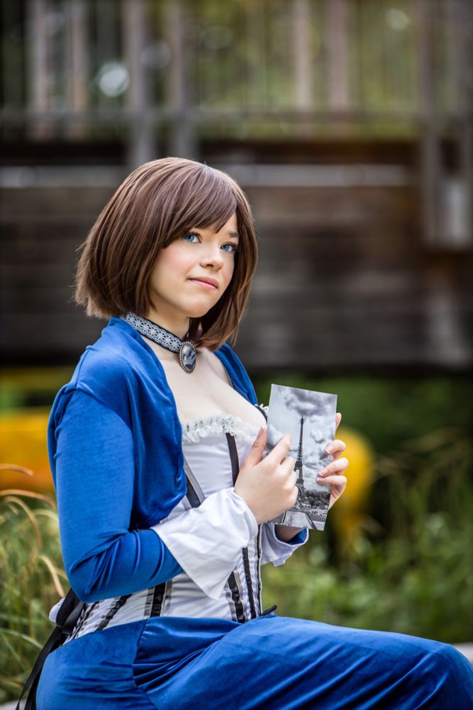 Eine junge Frau mit kurzen braunen Haaren und strahlend blauen Augen sitzt im Freien und hält eine schwarz-weiße Fotografie des Eiffelturms in den Händen. Sie trägt ein aufwendig gestaltetes blaues und weißes Cosplay-Kostüm mit viktorianischen Elementen, darunter ein elegantes Mieder, eine Halskette mit einem Medaillon und lange Ärmel mit Spitzenbesatz. Ihr sanfter Blick ist verträumt, als würde sie von einer fernen Welt oder einer anderen Zeit träumen. Im Hintergrund ist eine hölzerne Brücke mit leicht verschwommenem Grün zu sehen.