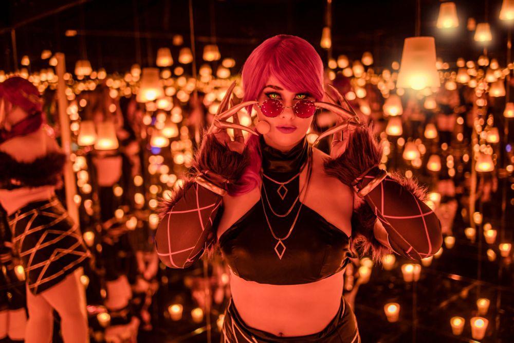 Eine Person mit leuchtend lila Haaren steht in einem Raum voller Spiegel und schwebender, warm leuchtender Lichter. Sie trägt ein schwarzes, glänzendes Crop-Top mit goldenen Ketten und geometrischen Anhängern, kombiniert mit langen, dunklen Armstulpen mit rautenförmigen Mustern und flauschigen Fellbesätzen. An den Fingern trägt sie lange, metallische Klauen. Eine runde, pink getönte Sonnenbrille sitzt auf ihrer Nase, die sie leicht mit den Klauen anhebt. Ihr intensiver Blick ist auf die Kamera gerichtet, während die unzähligen Spiegelbilder um sie herum eine magische, fast hypnotische Atmosphäre schaffen.