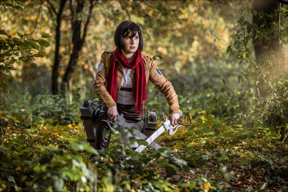 Eine Cosplayerin verkörpert Mikasa Ackerman aus der Anime- und Manga-Serie „Attack on Titan“. Sie trägt die typische Militäruniform des Aufklärungstrupps: eine braune Jacke mit dem Flügel-der-Freiheit-Emblem, weiße Hose, rote Schärpe und ihren charakteristischen roten Schal. Sie kniet im herbstlich gefärbten Wald, hält zwei Klingen der 3D-Manöverausrüstung in den Händen und wirkt kampfbereit und fokussiert.