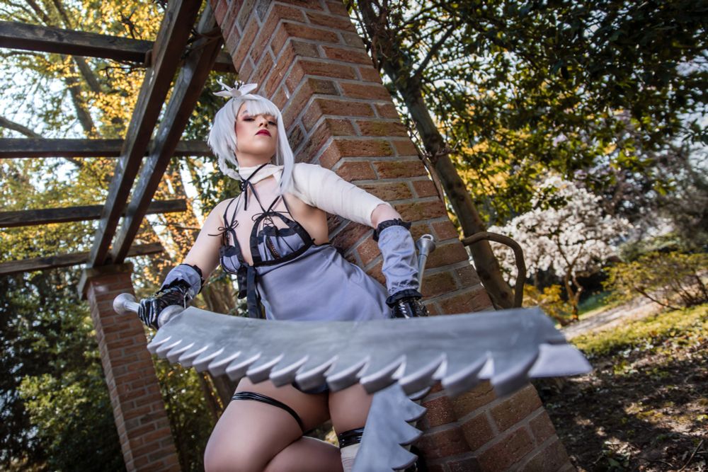Eine Person im Cosplay mit weiß-silberner Perücke und einem fantasievoll gestalteten Outfit steht kämpferisch an eine Ziegelmauer gelehnt. Sie trägt ein asymmetrisches Kleid mit Riemen und Bandagen am Arm. In der Hand hält sie ein großes, gezacktes Schwert, das kraftvoll nach vorn ragt. Der Hintergrund zeigt eine Gartenlandschaft mit blühenden Bäumen und warmem Licht, das durch das Laub fällt.