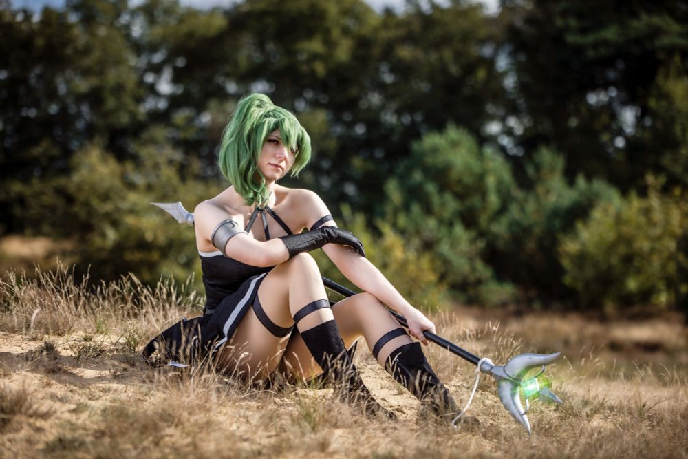 Eine Cosplayerin mit leuchtend grüner Perücke sitzt entspannt auf einer sandigen Wiese, umgeben von Natur und Bäumen. Sie trägt ein schwarzes, schulterfreies Kostüm mit silbernen Akzenten, fingerlose Handschuhe und hohe Stiefel mit Bändern. In ihrer Hand hält sie eine große, futuristisch anmutende Waffe mit einer leuchtend grünen Kristallspitze. Ihr Blick ist ruhig und fokussiert, während sie in die Ferne schaut. Die warme Sonne wirft ein sanftes Licht auf die Szene und verleiht ihr eine magische, fast märchenhafte Atmosphäre.