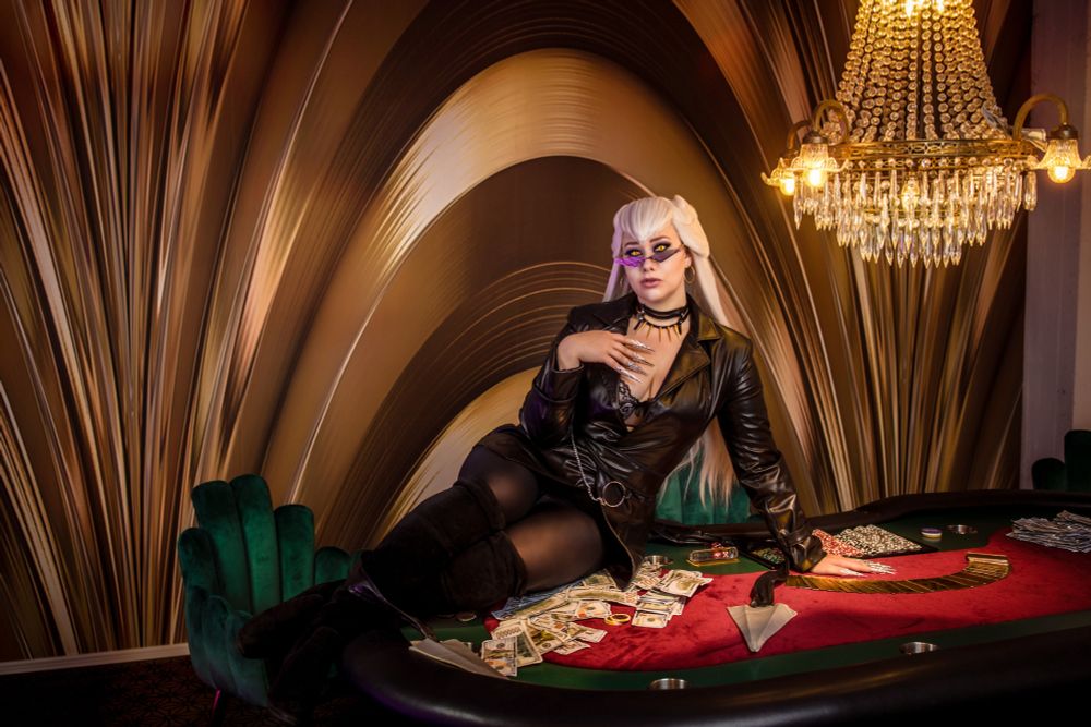 Eine stylische Person mit langen, platinblonden Haaren und kleinen violetten Sonnenbrillen sitzt an einem roten Pokertisch in einem opulenten Kasinoambiente. Sie trägt eine schwarze Lederjacke und dunkle Kleidung und posiert selbstbewusst, während Geld über den Tisch verstreut liegt. Der Hintergrund zeigt elegante, goldfarbene geschwungene Wandpaneele, und ein Kristallleuchter ist in der oberen rechten Ecke sichtbar. In der Nähe steht ein grüner Samtsessel. Die gesamte Szene strahlt eine luxuriöse, hochwertige Glücksspielatmosphäre aus.