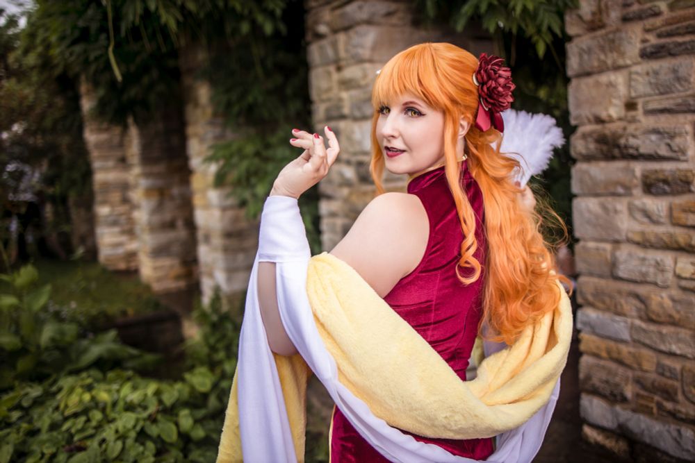 Eine Cosplayerin stellt Nami aus One Piece dar, in ihrer eleganten Cheongsam-Version. Sie trägt ein dunkelrotes, figurbetontes Kleid im chinesischen Stil mit freiem Rücken und Schultern. Ihr langes, orangefarbenes Haar ist in Locken gestylt und mit einer großen roten Blume geschmückt. Über ihre Arme liegt ein weiß-gelber, weicher Umhang. In der Hand hält sie einen weißen Fächer, während sie sich mit einem charmanten Lächeln über die Schulter zur Kamera dreht. Im Hintergrund ist eine mit Efeu bewachsene Steinmauer zu sehen.