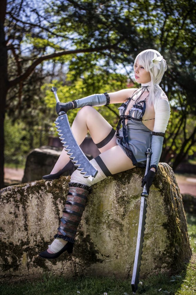 Eine Person cosplayt Kaine aus dem Videospiel NieR Replicant. Sie sitzt auf einem bemoosten Felsen in einem sonnigen Park mit grüner Kulisse. Das Outfit besteht aus einem freizügigen, grau-blauen Kostüm mit asymmetrischen Elementen, Bandagen, Armstulpen und dekorativer Beinschiene. Die Cosplayerin trägt eine silberweiße Perücke mit einer Blume im Haar und hält Kaine’s ikonisches gezacktes Schwert locker in einer Hand. Der Gesichtsausdruck ist ernst und fokussiert.