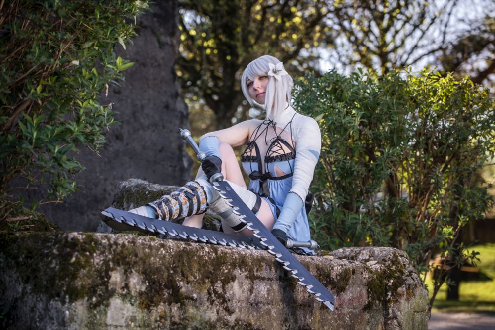 Person in aufwendigem Cosplay sitzt auf einer bemoosten Steinmauer im Freien, umgeben von grünen Büschen und Bäumen. Das Kostüm besteht aus einem kurzen, blaugrauen Kleid mit Cutouts, langen Armstulpen und aufwändigen Beinschienen. Die Person hat silberweißes Haar mit einer großen Blume als Haarschmuck. In der Hand hält sie ein großes, gezacktes Schwert. Die Pose ist selbstbewusst und entspannt, der Blick direkt zur Kamera gerichtet. Die Szene wirkt märchenhaft und leicht mystisch.
