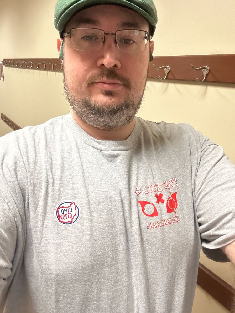Scruffily bearded man wearing a grey Fractured Fox shirt and green flat cap with an Ohio I Voted sticker.