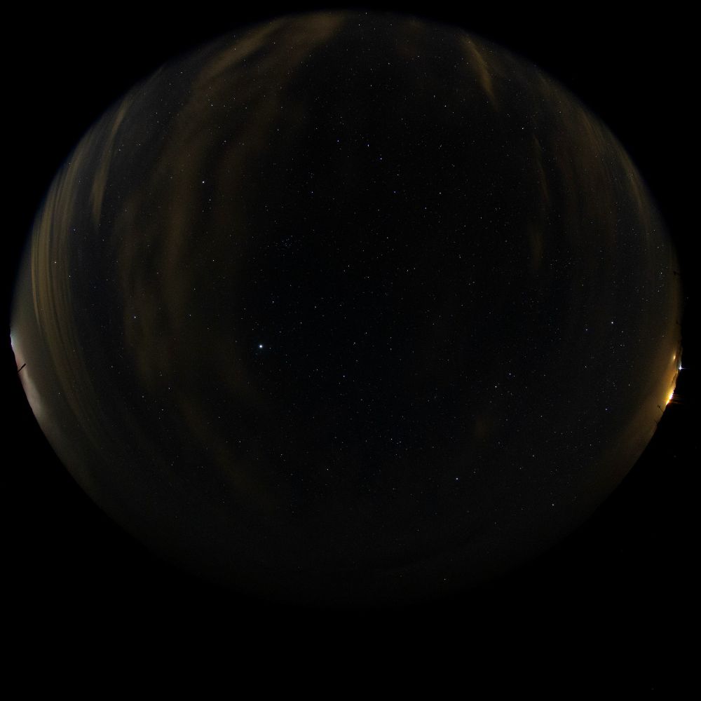 A fisheye photo of the sky, replete with light yellow-cast clouds and severe skyglow to the right of the image.