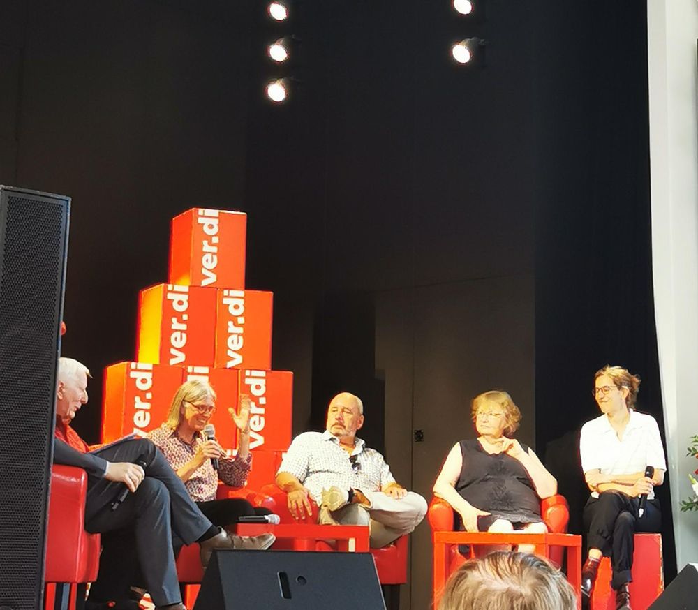 5 Kolleg*innen in roten Stühlen in einem Halbkreis Auf einem Podium. 
