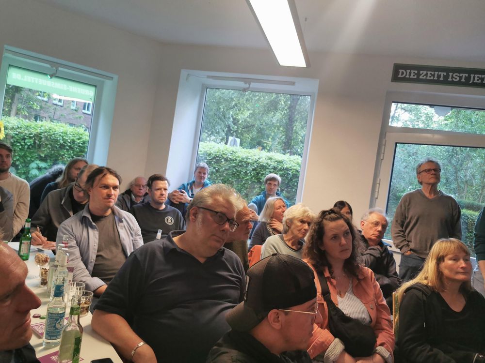 Blick auf ca. 25 dicht gedrängt sitzende Menschen, die einem Referenten lauschen, der nicht zu sehen ist. 