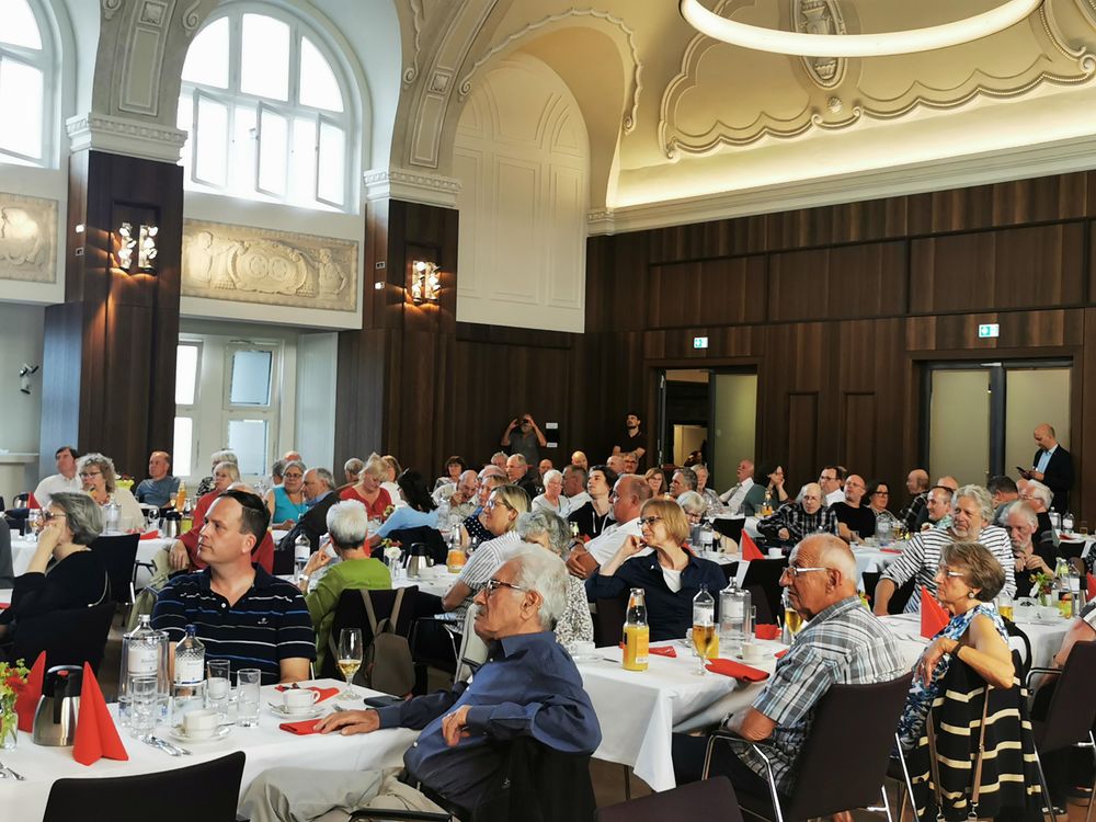Blick auf drei Tischreihen voll besetzt mit Menschen, die in Richtung Bühne gucken. 