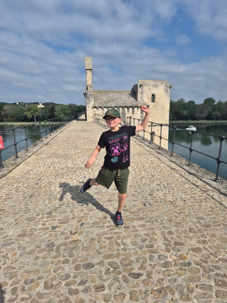 My beautiful son, Bertie button, dancing on the bridge in Avignon, "sur le pont d'Avignon" with joy and freedom. The sun is shining and there are white clouds speckled through the sky. Bertie is 10, wearing a Minecraft t-shirt, green shorts and a Titanic Belfast hat, and looks every bit the delightful, big hearted pickle that he is 
