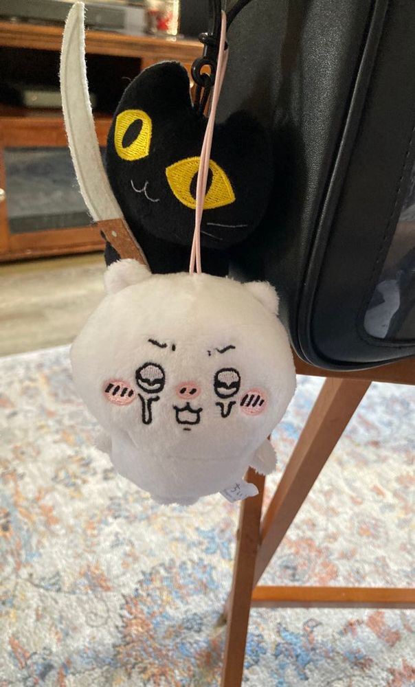 A crying chiikawa plush with a black cat plush holding a knife menacingly behind him. They are both attached to a black purse.