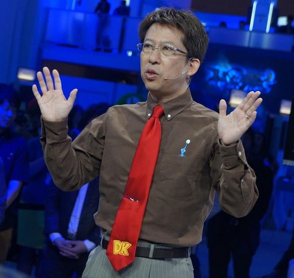 picture of Kensuke Tanabe from Nintendo's 2013 E3 booth in a brown shirt, sporting a DK tie with a Madonna headset and glasses, 90° arms with open hands poking skyward, gaze to the off-camera horizon ...essentially matching his son, Sylux.