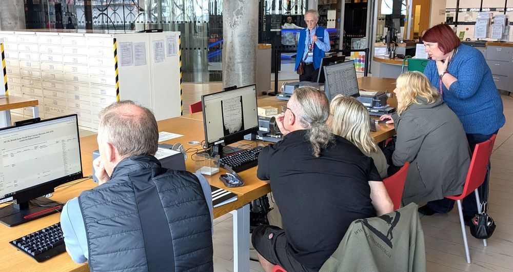 Photo 1 -  Family History Taster session. Four people with their backs to the camera sat in a row looking at computer screens. A female figure at the end of the row on the far right of the photo is standing and bent slightly over the table providing guidance. A male figure to the left of the group stands and observes. 