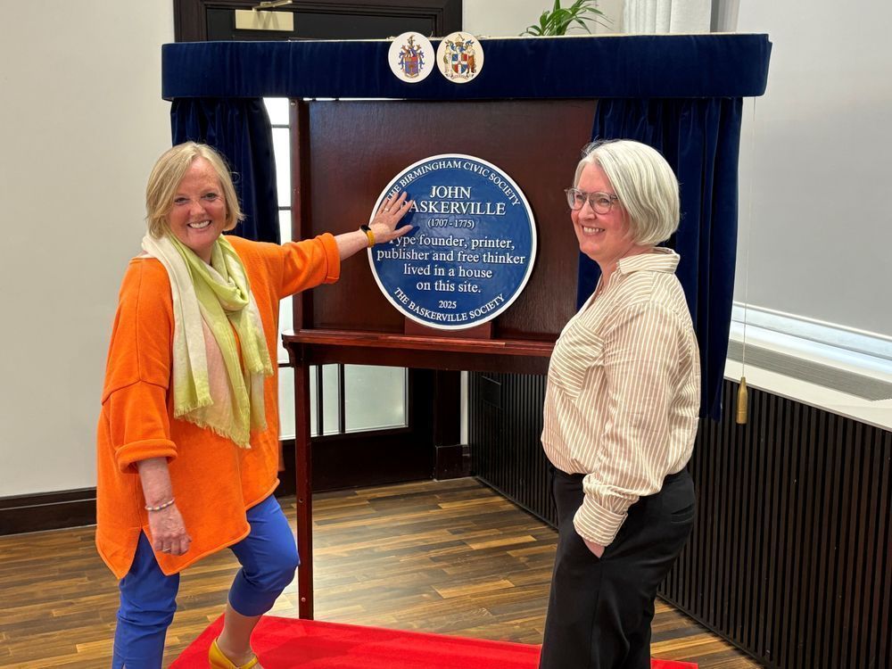 A room with French windows in the background. A stand with a blue civic society plague on it  -  the plague is dedicated to John Baskerville 1707 – 1775, type founder, printer, etc. Two female figures are stood on either side of the plaque and stand. The figure on the left appears to be touching the plague. Both figures are smiling. 