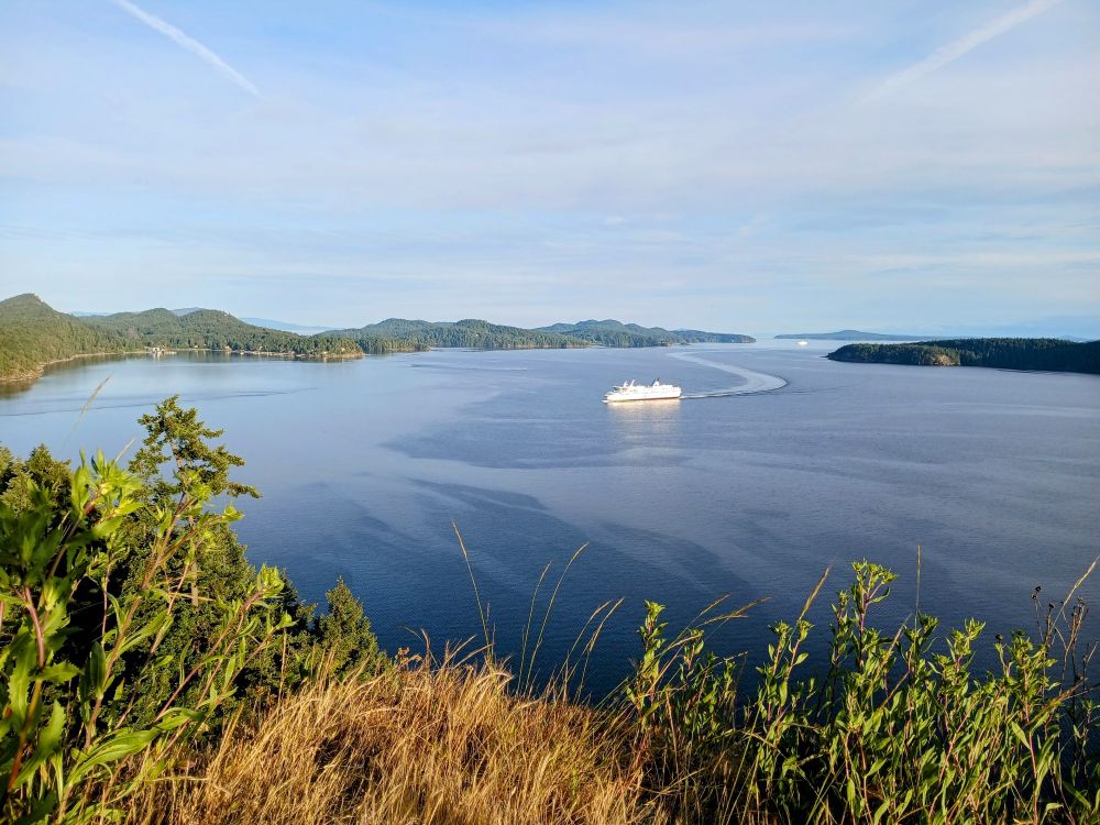View from a high point on Galiano Island. Other islands are visible, the sea is calm, and a ferry is making its way to the island, leaving a gentle wake trail behind it. Another ship is in the far distance.