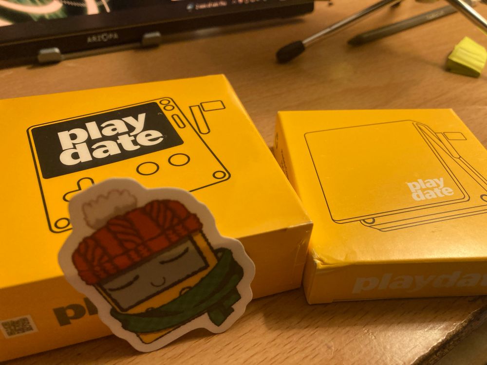 Package of a Playdate on the left, the Playdate Case on the right, and a sticker of a Playdate with a toque and a scarf chilling