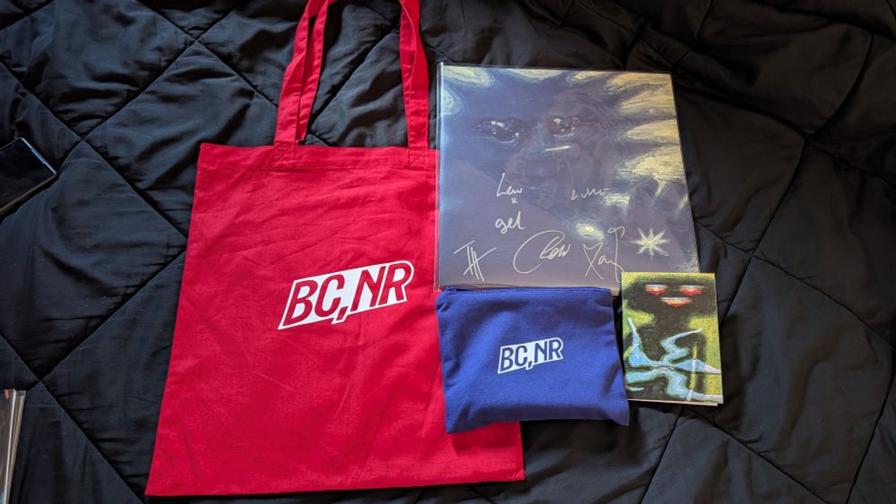 red tote bag with BC,NR logo, signed Forever Howlong vinyl album, smaller blue zipup bag with BC,NR logo, and small notebook with artwork cover.