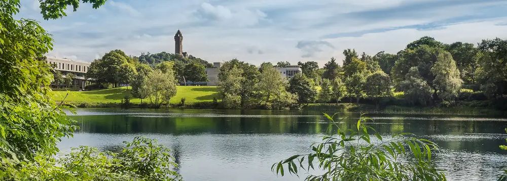 This photograph of Stirling campus shows part of the campus lake with green grass, trees and the outline of the Wallace monument on a wee hill in the background.