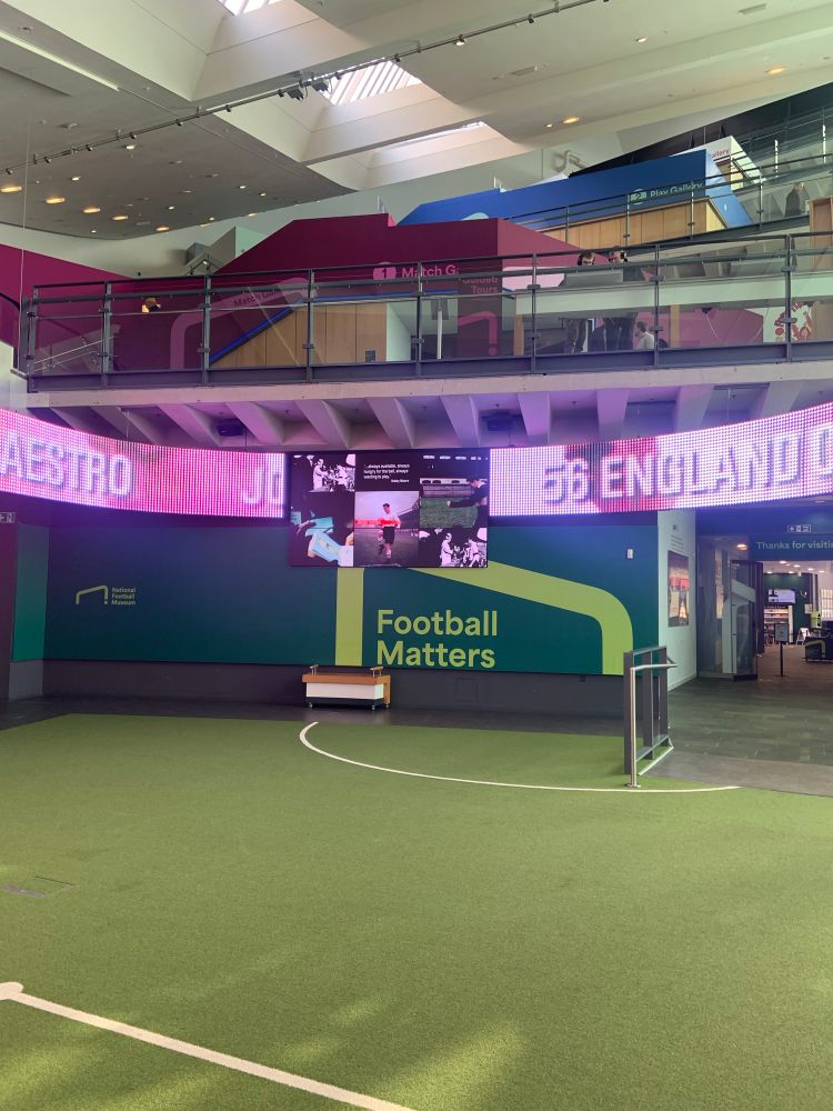 Ground floor main room of museum with an astroturf carpet with soccer field markings. The back wall says “Football matters” inside a goalpost. Electronic boards line the top of the room