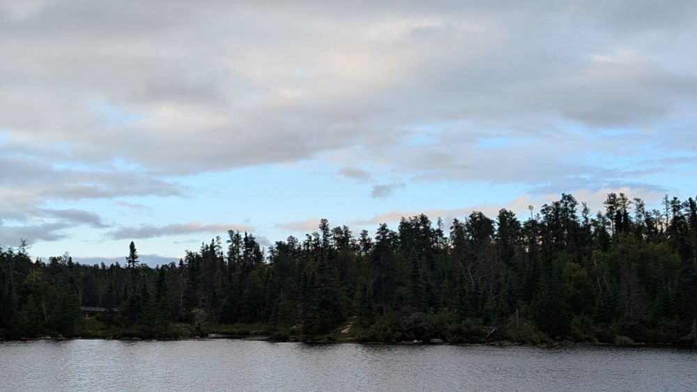 A view across the calm water of a lake to a shoreline lined with a dense, dark evergreen forest under a cloudy sky with patches of blue.
