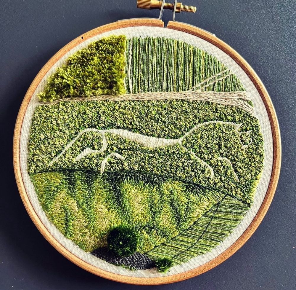 A circular embroidery in a wooden hoop of the White Horse using various stitches including hundreds of French knots for the main grass, long and short for the folds of the hill below the horse, and turkey tail for a field and scattered trees. 