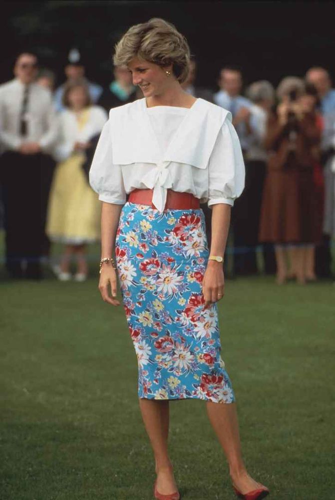 Une photo de Lady Diana en 1985 vétue d'un jupe moulante mi-longue bleue fleuri taille haute, serrée à la taille par une large ceinture en cuir marron, avec une blouse blanche bouffante à col marin manches mi-longues rentrée dedans.