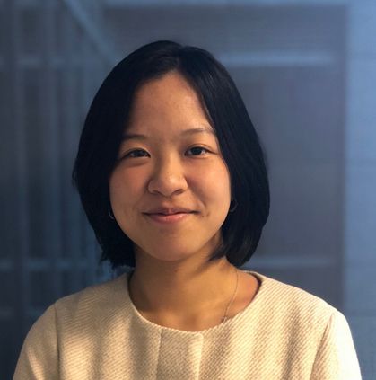 Photographic portrait of Anin Luo facing the camera, wearing a cream suit jacket, a small silver necklace, and black bobbed hair. Anin is smiling while appearing in front of a photograph of a blue building.