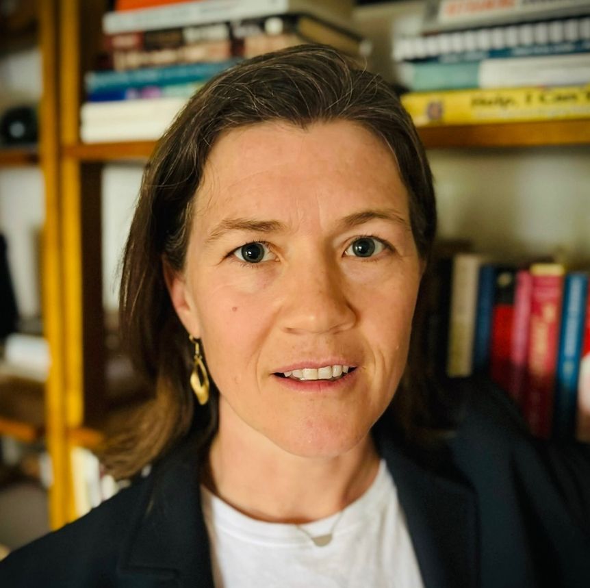 Photograph of Emily Yates-Doerr next to a bookshelf stacked with books, Emily is wearing a white t-shirt, and black jacket with shoulder length brown hair, large golden hoop earring and a semi-circle gold necklace. 