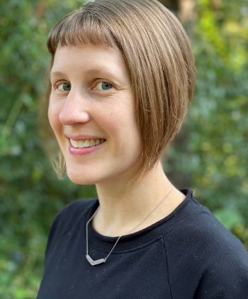 Photographic portrait of Kim De Wolff facing the camera side-on. Kim is wearing a dark navy-black sweater and a gun-metal silver v-shaped necklace. Kim is smiling at the camera with brown bobbed hair, with a green background pictured outdoors.