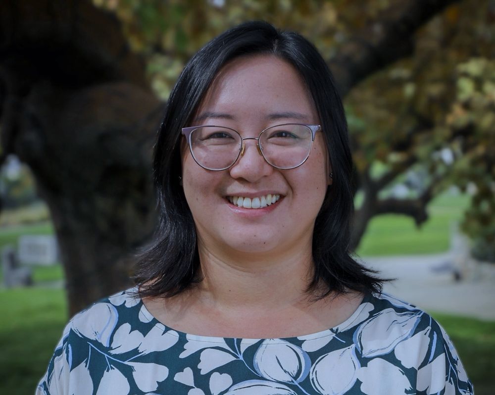 A photograph of Lisa Yin Han smiling at the camera, pictured outside in front of a tree and a grassy background, and is wearing metal glasses and a botanical printed top.