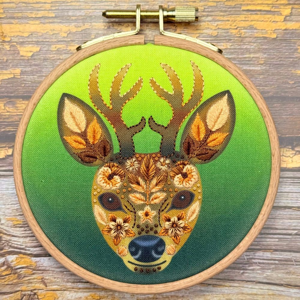 Floral‑patterned stag head stitched in warm browns, gold and cream threads, set in a wooden embroidery hoop against a gradient green fabric background