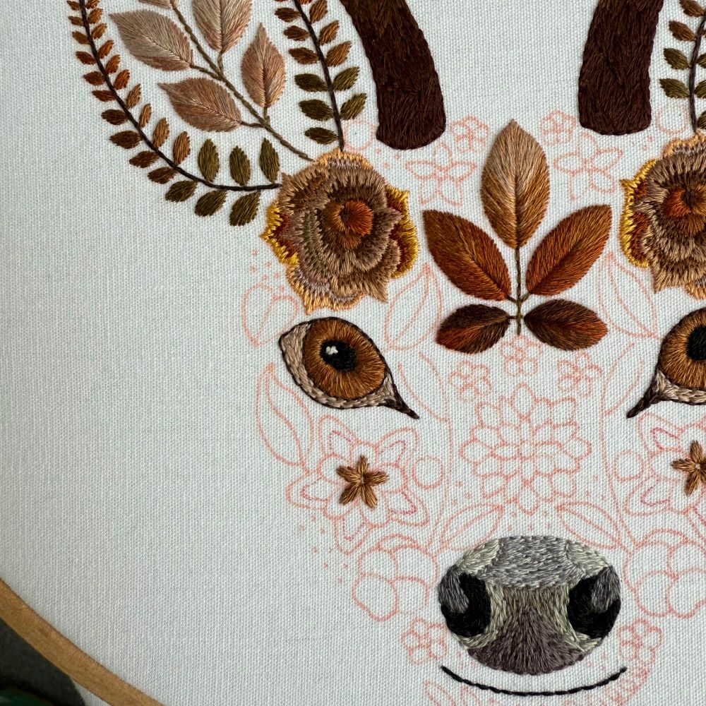 Photo of an embroidered stag. The stags face is created using flowers and leaves. The antlers, ears, eyes, nose and mouth are stitched in brown threads. The areas of the stags face that are not yet stitched are marked out in red fabric pen. The fabric is white. 