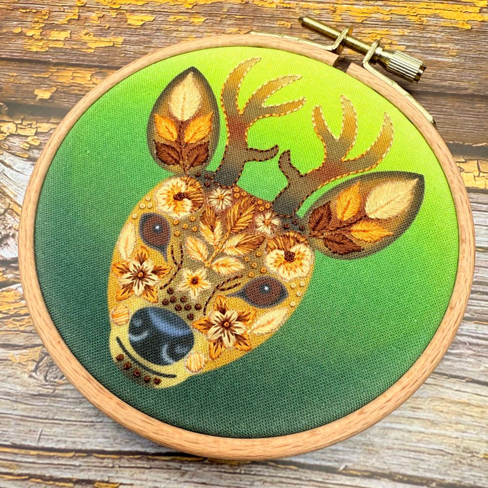 Hand‑stitched stag silhouette with floral embroidery details—leaves and flowers in autumn tones framed in round wooden hoop on green canvas