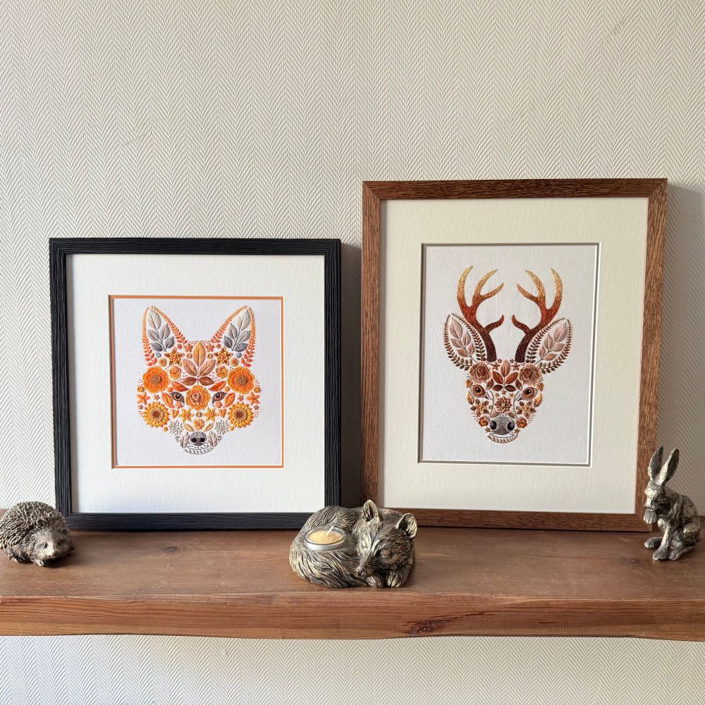 Two framed embroidered artworks displayed on a wooden shelf against a light herringbone-patterned wall. The left frame features a fox’s face made from orange, yellow, and brown floral and leaf motifs, with a black frame and orange inner border. The right frame shows a stag’s head with brown and golden antlers, also created from floral and leaf embroidery, in a wooden frame with cream mount. Small animal figurines, including a hedgehog, sleeping fox candle holder, and hare decorate the shelf.