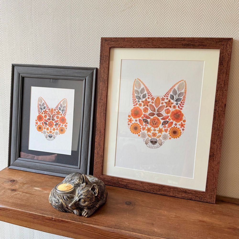 Photo of two photo frames on a brown wooden shelf. Both frames feature the same artwork, a foxes face created using flowers and leaves. The frame on the left is grey with a black boarder surrounding the artwork. The frame on the right is brown with a cream boarder surrounding the artwork. In front of the frames is a candle holder that is shaped like a fox that is curled up asleep. The wall behind the frames is a cream colour. 