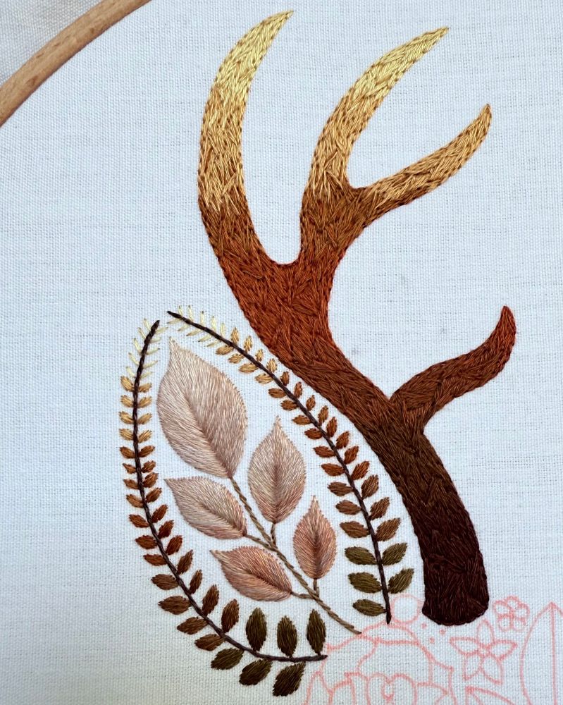 Photo of embroidery. Close up of an ear and antler on a floral stag design. The ear is stitched using leaf designs. The antler is densely stitching using various shades of brown. The antler has an ombré effect with lighter shades at the top and darker shades at the bottom. The embroidery is stitched on white fabric. 