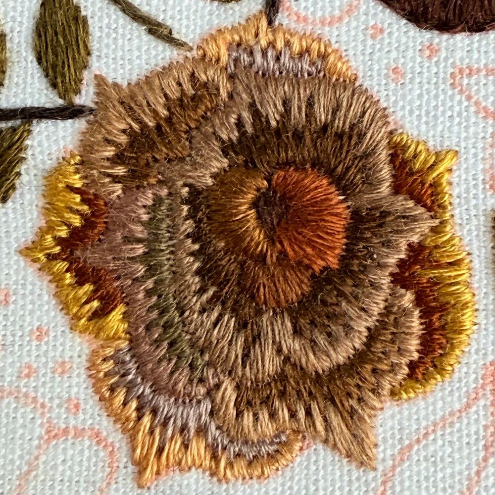 Close up photo of an embroidered rose. The rose is stitched using various shades of brown. The rose is stitched onto white fabric. 