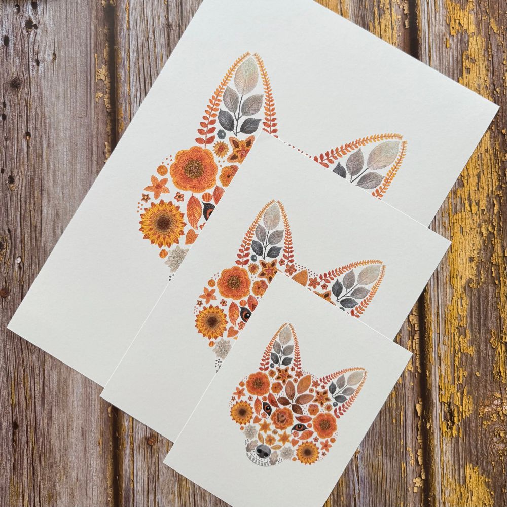 A photo showing 3 floral fox art prints. The art prints are of the same image but are in 3 different sizes. The design is of a foxes face created from flowers and leaves. The largest print is at the back with the smallest at the front of the stack of art prints. They are displayed diagonally. The background is brown wood. 