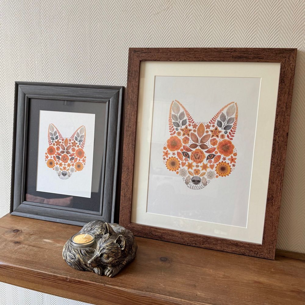 Two framed artworks of a floral fox art print. The frame of the left is grey with a black boarder surrounding the art print. The frame on the right is brown wood with a cream boarder surrounding the art print. Both art prints are of a foxes face created using flowers and leaves. They are sat on a wooden shelf. The wall behind is cream coloured. In front of the frames is a candle holder in the shape of a fox curled up sleeping. 