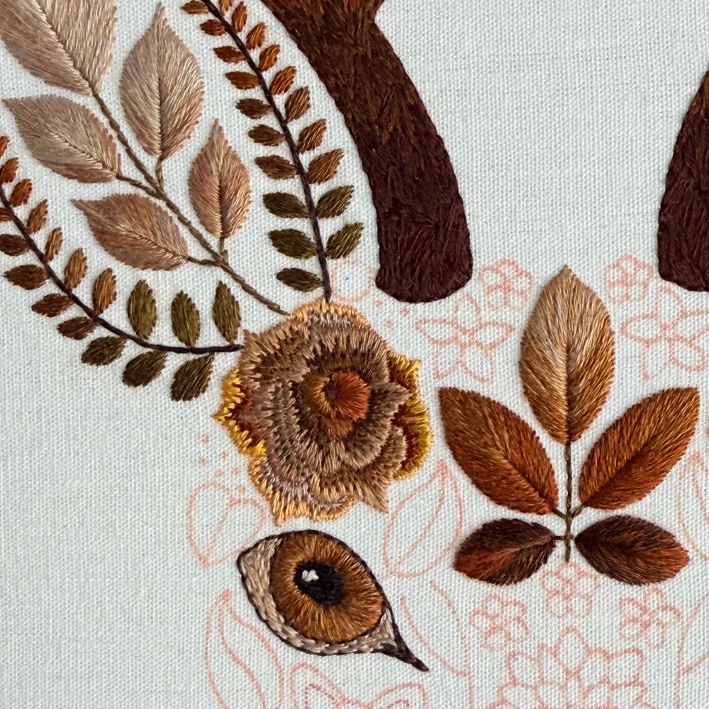 Photo of part of an embroidered stag. The top left part of the head is visible. The eye, ear and antler are stitched using different shades of brown thread. There is a brown rose and leaf cluster stitched on the head of the stag. The embroidery is on white fabric. 