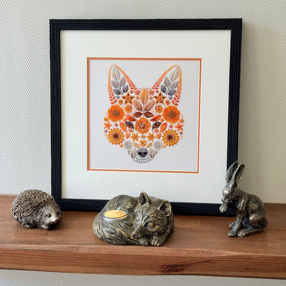 Photo of a hand embroidery of a foxes face created using flowers and leaves. The artwork is framed and sitting on a brown wooden shelf. The frame is black and made from textured wood. It is a square shape. There is a cream boarder around the artwork with a smaller boarder of orange. Sat in front of the frame and 3 bronze metal ornaments, a hedgehog, a fox and a hare. The wall behind the artwork is cream. 