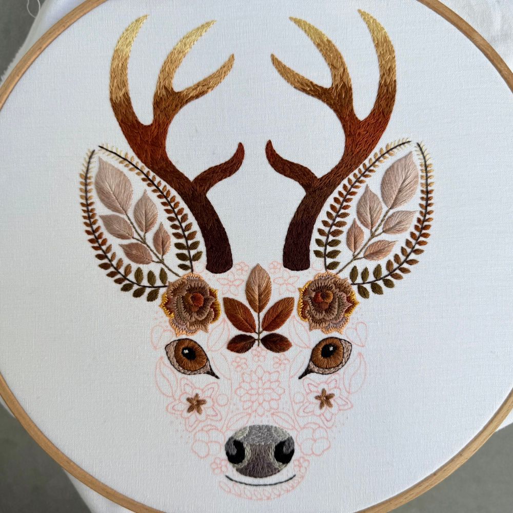 Photo of white fabric in a wooden embroidery hoop. The design on the fabric is of a stags face created using flowers and leaves. The antlers, ears, eyes, nose and mouth are stitched in shades of brown thread. The stags face also has two brown roses and a leaf cluster stitched. The rest of the face is marked out in red fabric pen waiting to be embroidered. 
