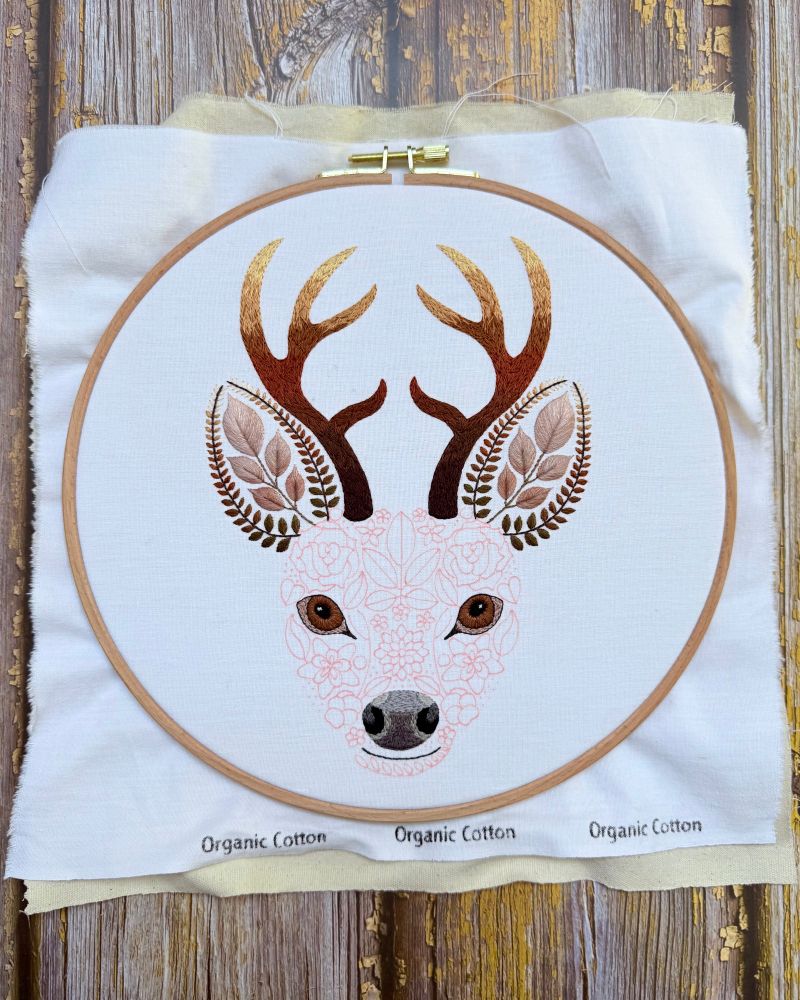 Photo of a stag embroidery. The design is created using flowers and leaves. Part of the stag has been stitched and the rest of the design is marked out in red fabric pen. The embroidery is on white fabric. The fabric is secured in a wooden embroidery hoop. 