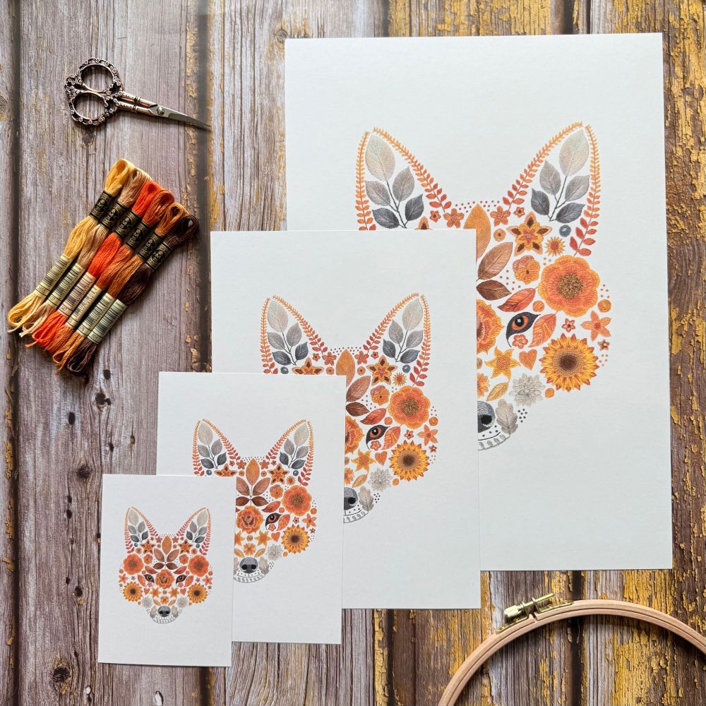 Photo of a 4 different sized floral fox art prints. The background of the photo has a wooden effect in shades of brown and orange. The art prints are off white and features a foxes face created with flowers and leaves. There is a large print with three smaller sized prints stacked in front. Next to the prints are some embroidery scissors, an embroidery hoop and six shades of thread varying from light yellow to dark brown.