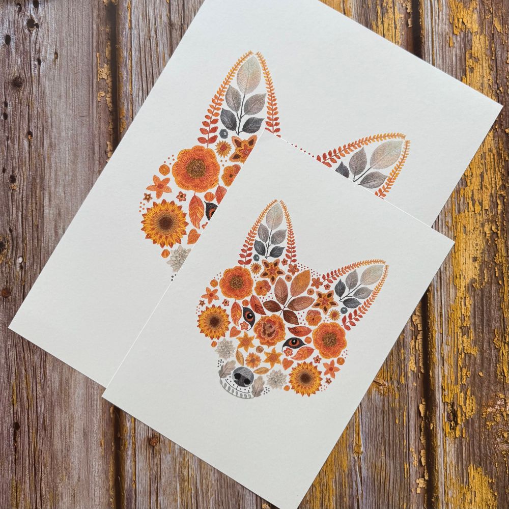 A photo showing 2 floral fox art prints. The art prints are of the same image but are in 2 different sizes. The design is of a foxes face created from flowers and leaves. The largest print is at the back with the smallest at the front. They are displayed diagonally. The background is brown wood. 
