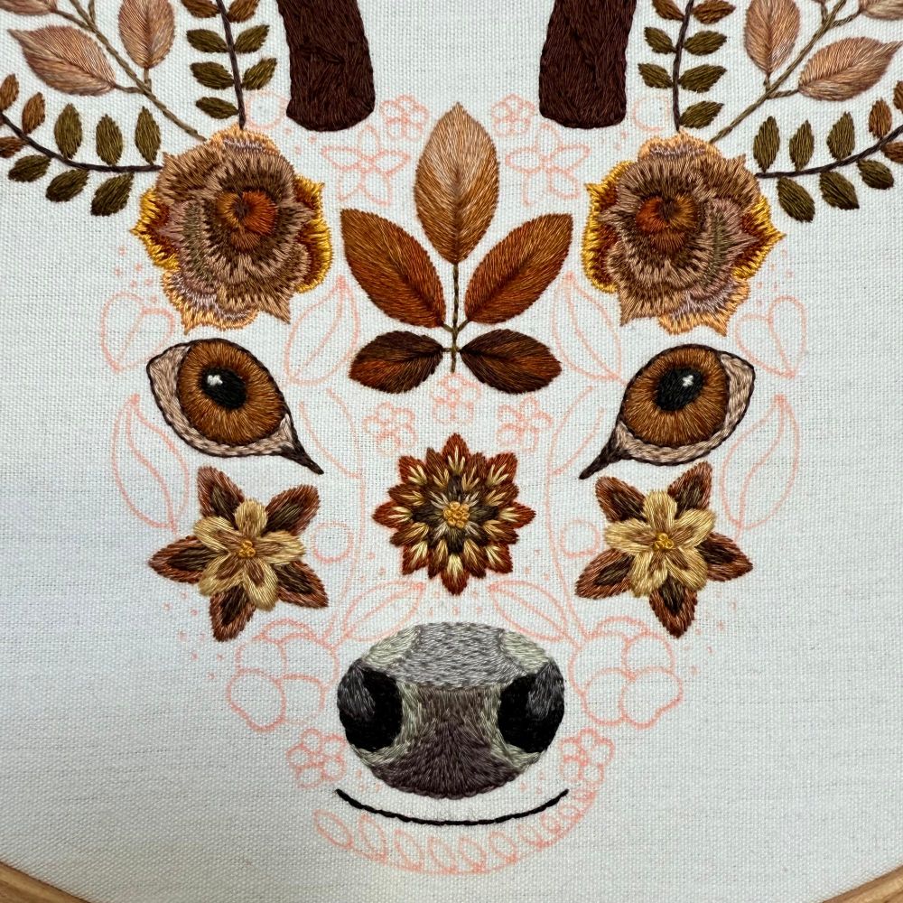 Photo of an embroidery of a stags face. The face is created using flowers and leaves. The photo is a close up of the face. The areas of embroidery are created using brown threads. The areas yet to be embroidered are marked out in red fabric pen. The fabric is white. 